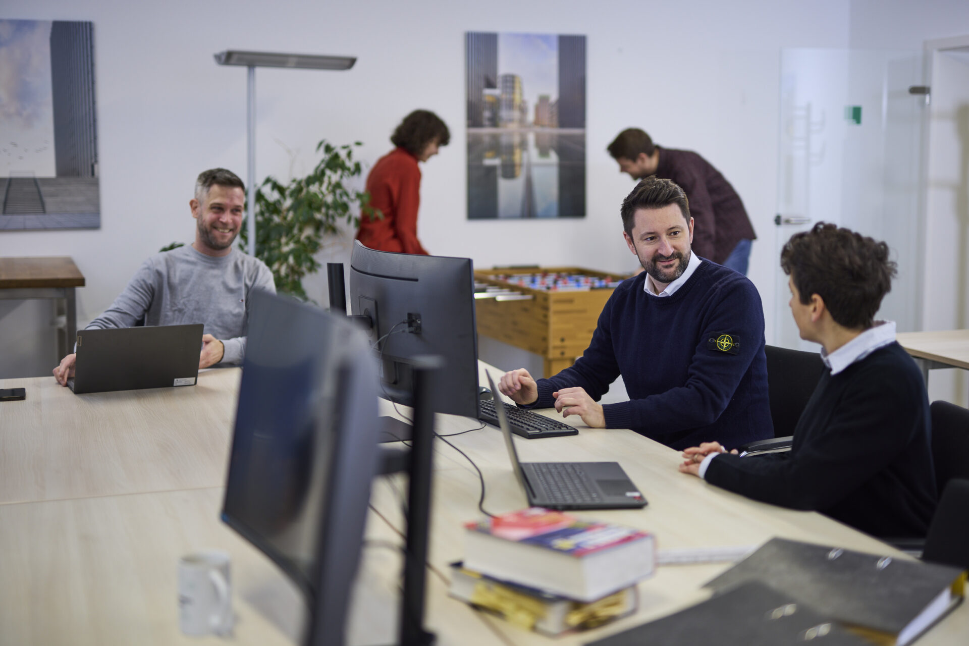 Mehrere Personen sitzen an einem Tisch mit Computern und unterhalten sich in einem modernen Büro mit Tischkicker im Hintergrund